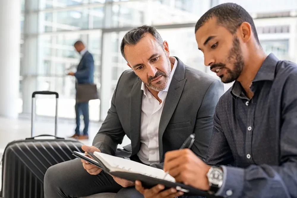 Two multiethnic businesspeople sitting at departure lounge at the airport discussing work while making notes in agenda. Indian b